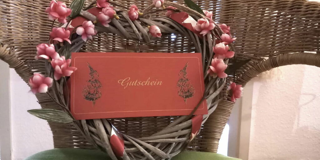 Ein Thai Massage Geschenkgutschein vor einem geflochtenen Herz mit Blumen als Symbol für ein schönes Valentinstagsgeschenk.