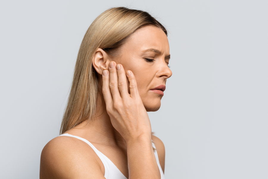 Eine mittelalte Frau mit langen blonden Haaren und geschlossenen Augen hält sich das Ohr, da sie unter Tinnitus leidet.